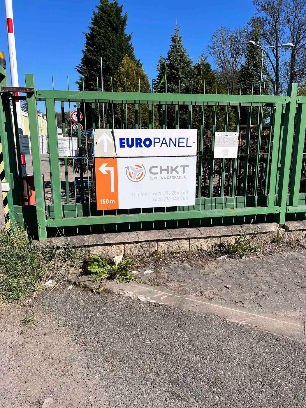 Směrová cedule CHKT tepelná čerpadla na zelené bráně u vjezdu do areálu, šipka vlevo a údaj 150 m